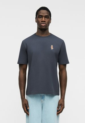 Man draagt een donkergrijs T-shirt met korte mouwen en een kleine geborduurde vos op de linkerborst, gecombineerd met lichtblauwe spijkerbroek, tegen een effen achtergrond.