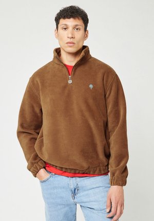 Man wearing a brown corduroy half-zip pullover with elastic cuffs and hem, red shirt underneath, and light blue jeans, standing with one hand in pocket.