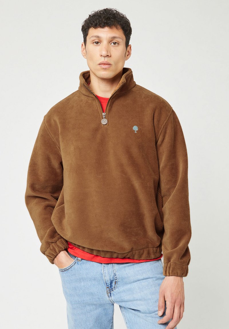 Hombre vestido con un pulóver de media cremallera de pana marrón con puños y dobladillo elásticos, una camisa roja debajo y jeans de color azul claro, de pie con una mano en el bolsillo.