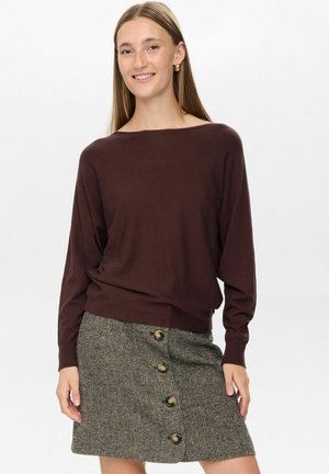 Maglione marrone, oversize, con scollo a spalle scoperte, abbinato a una gonna con motivo a spina di pesce e bottoni dorati.