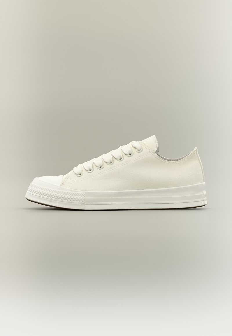 Λευκό χαμηλό αθλητικό παπούτσι από καμβά με λευκή ελαστική σόλα, προστατευτικό στη μύτη και λευκά κορδόνια, εμφανίζεται από το πλάι σε ουδέτερο φόντο.