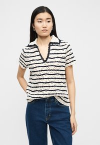 Νεαρή γυναίκα με μακριά σκούρα μαλλιά, που φοράει ένα μπεζ και μαύρο ριγέ πλεκτό τοπ με κοντά μανίκια και μπλε τζιν, στέκεται μπροστά σε απλό φόντο.