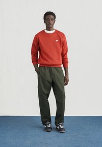 Sweatshirt vermelha com um pequeno logotipo branco, calças cargo verdes e ténis pretos. Modelo está em pé num tapete azul contra um fundo claro.