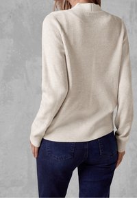 Žena v béžovém žebrovaném svetru a tmavě modrých džínách, stojící zády k fotoaparátu proti šedé strukturované zdi.