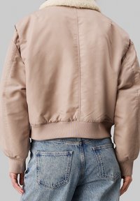 Lichtbeige bomberjack met een zachte witte kraag, geribde manchetten en tailleband, en een gladde stofafwerking, gedragen met vervaagde blauwe jeans.