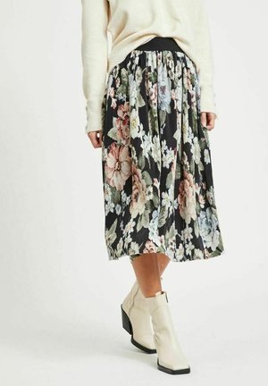 Kvinde iført plisseret blomsterprintet midi-nederdel med sort baggrund, cremefarvet sweater og hvide ankelstøvler, stående mod en ensartet lys baggrund.