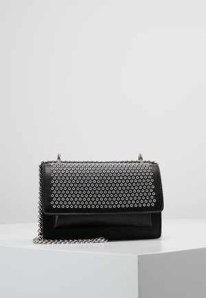 Czarna skórzana torba na ramię z srebrnymi nitami w kształcie oczek na klapie oraz srebrnym łańcuszkiem jako paskiem, na białej powierzchni.