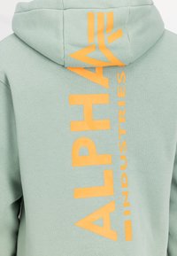 Mentazöld kapucnis pulóver, amelynek bal oldalán merőlegesen, nagy narancssárga betűkkel olvasható az "ALPHA INDUSTRIES" felirat. Puha anyag, kenguru zseb.