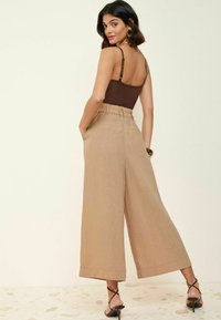 Beige, weit geschnittene Hose aus strukturierter Stoff mit elastischem Bund und seitlichen Taschen, kombiniert mit einem braunen Body und schwarzen Riemchen-Heels.
