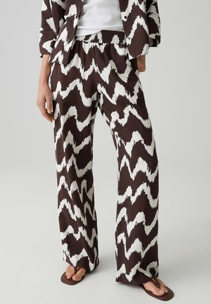 Vrouw draagt zwart-wit zigzagpatroon wijde broek, bijpassend jasje, wit topje en bruine sandalen, staand op een lichte achtergrond.