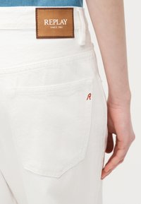 Πίσω τσέπη λευκού τζιν με μικρή κόκκινη ραφή "R" και καφέ δερμάτινο ένθετο με την επιγραφή "REPLAY SINCE 1981" κοντά στη μέση μπλε πουκαμίσου.