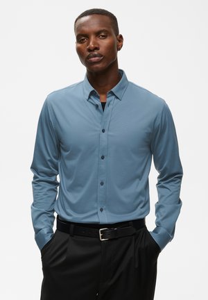 Homme portant une chemise bleu clair boutonnée et un pantalon noir, les mains dans les poches, sur un fond blanc uni.