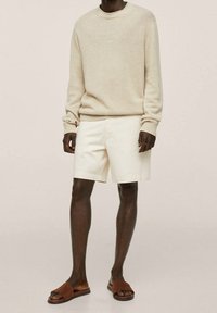 Beiger Strickpullover mit Rundhalsausschnitt, kombiniert mit off-white Shorts. Braune Wildledersandalen. Sauberes Design, legerer Schnitt, texturierter Stoff.