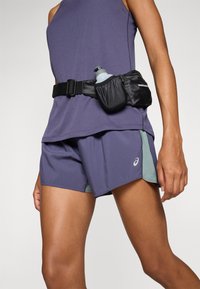 Top deportivo morado combinado con pantalones cortos a juego que tienen paneles laterales verdes. Una riñonera negra sostiene una botella de agua gris.