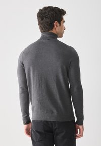 Hombre con cabello rizado oscuro, vistiendo un suéter gris ajustado de cuello alto y jeans negros, visto desde atrás contra un fondo blanco liso.