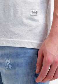 Šedé tričko s krátkým rukávem vyrobené z texturované látky s nášivkou G-Star. V kombinaci s světle modrými džínami s efektem opotřebení. Viditelné detaily šití.