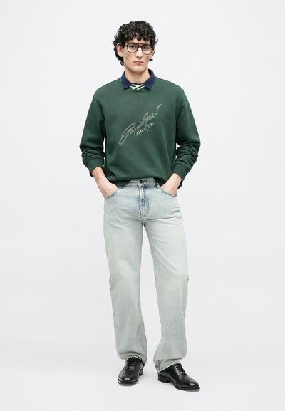 Πράσινο φούτερ με χαρακτηριστικό σχέδιο, μπλε πουκάμισο με γιακά από κάτω, ανοιχτόχρωμο τζιν, και μαύρα παπούτσια. Το μοντέλο στέκεται με τα χέρια στις τσέπες.