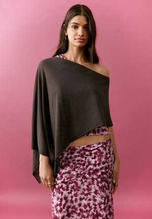 Jonge vrouw met lang bruin haar, een zwarte off-shoulder poncho dragend over een paars bloemen twee-delig outfit, tegen een effen roze achtergrond.