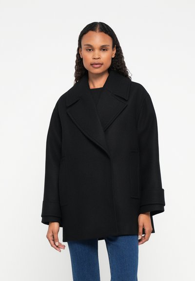 Femme portant un manteau noir surdimensionné avec de larges revers et un jean bleu, se tenant devant un fond blanc uni.