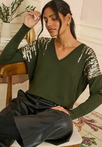 Olīvzaļa džemperis ar V-veida kakla izgriezumu un garām piedurknēm, rotāts ar zeltainām piešuvēm plecos. Apvienots ar melniem mākslīgās ādas biksēm.