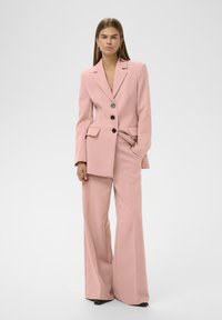 Frau, die in einem weit geschnittenen pinken Anzug mit schwarzen Knöpfen steht, eine Hand in der Tasche, mit Creolen, neutralem Ausdruck vor einem weißen Hintergrund.