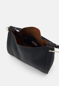 Sac à main en cuir noir avec ouverture supérieure montrant l'intérieur marron et une poche zippée à l'intérieur, sur fond blanc.