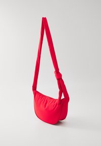 Borsa a spalla di stoffa rossa con forma arrotondata, caratterizzata da una tracolla spessa e annodata e un design morbido e strutturato. Testura liscia con dettagli minimi.