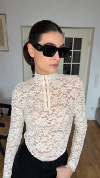 Woman with dark hair in low bun wears black Prada sunglasses and a white floral lace high-neck top, standing indoors near a glass-paneled door.