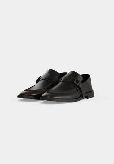 Paire de mocassins en cuir marron foncé sans lacets avec boucle latérale et petit talon sur fond clair uni.