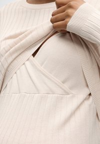 Personne soulevant un pull côtelé beige clair pour révéler un soutien-gorge d’allaitement côtelé beige assorti en dessous.