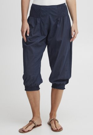 Femme portant un pantalon court bleu marine avec taille et poignets élastiques, associé à des sandales plates marron.