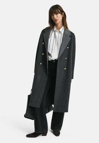 Grijze houndstooth jas met een dubbelknoopontwerp, gouden knopen en een brede kraag, gecombineerd met een wit gestreept overhemd en zwarte broeken.