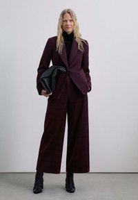 Traje de cuadros burdeos con pantalones de pierna ancha, blazer entallado y un bolso grande negro. Combinado con una blusa negra ajustada y botas de tobillo.