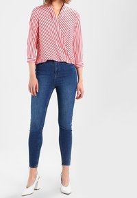 Rød og hvidstribet omslagbluse med trekvartærmer, parret med tætsiddende blå denimjeans og hvide hælede sandal. 