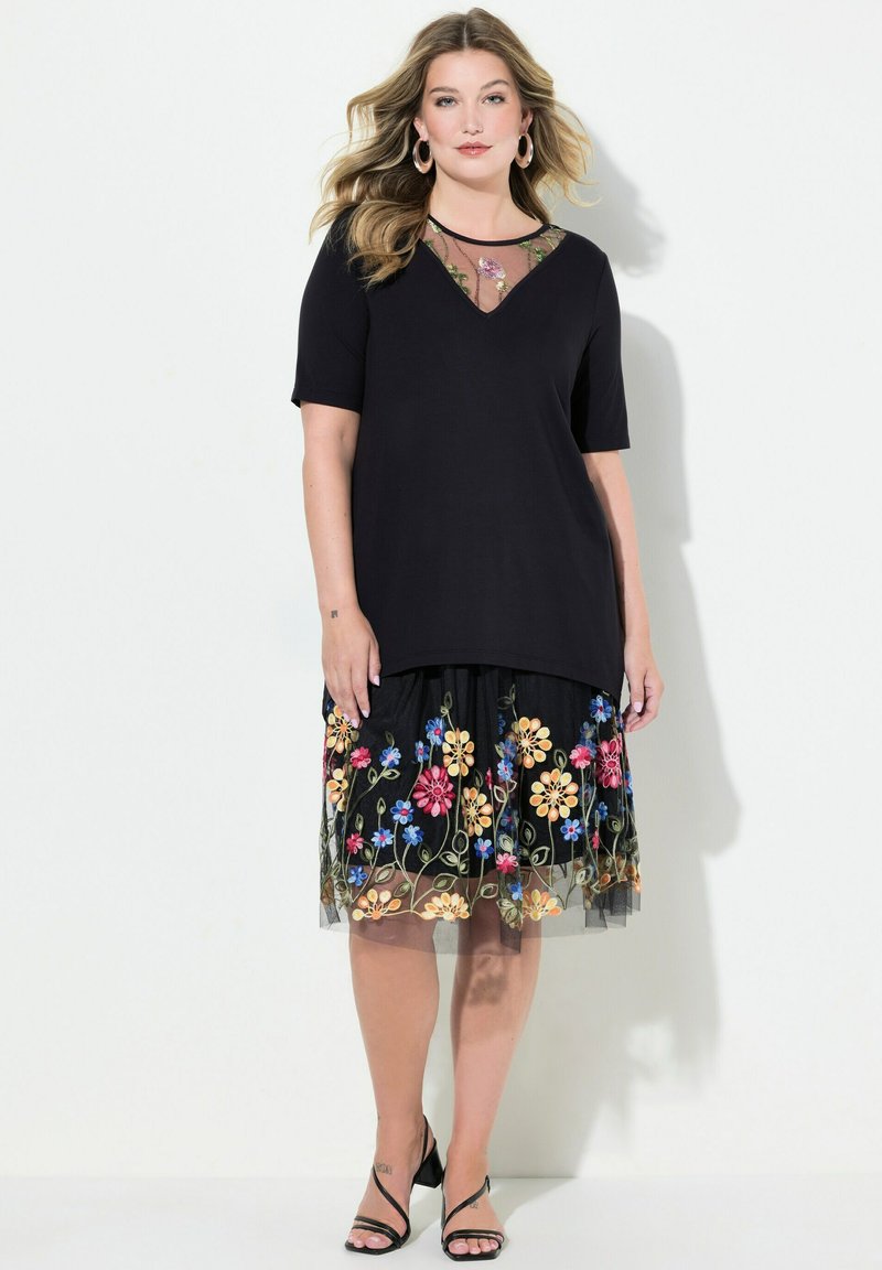Top negro con escote en V y mangas cortas, combinado con una falda negra con bordados florales coloridos y una superposición de tul. Tacones negros.