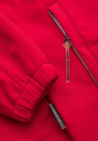 Gros plan d'une manche de veste rouge avec un poignet élastique et une poche zippée avec une languette étiquetée "ombre performance."