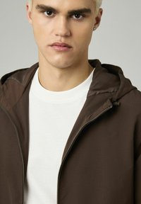 Veste à capuche marron avec une texture légère et une fermeture éclair, associée à une chemise blanche. La vue de près met en avant les détails du tissu et la coupe.