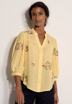 Frau trägt eine lockere gelbe Bluse mit 3/4-Ärmeln und bunter Blumenstickerei, kombiniert mit dunkler Hose vor einfarbigem Hintergrund.