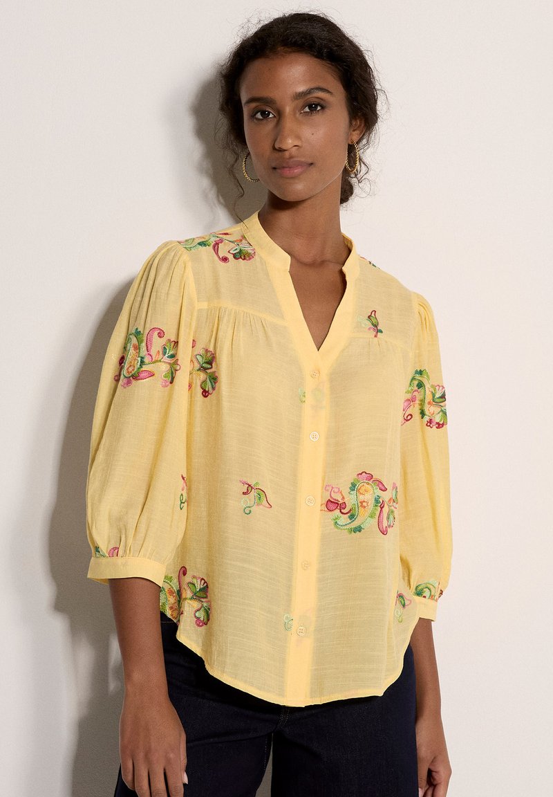 Mujer con blusa amarilla holgada de mangas tres cuartos y bordado floral colorido, combinada con pantalones oscuros sobre un fondo liso.