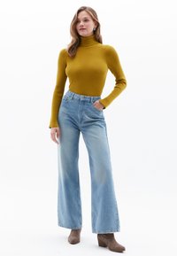 Musztardowy sweter z golfem o ryflowanym wzorze zestawiony z jasnoniebieskimi spodniami z wysokim stanem i szerokimi nogawkami oraz brązowymi botkami. Gładka tekstura, swobodny styl.