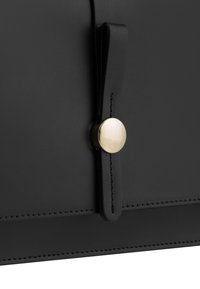 Dettaglio di una borsa in pelle nera con chiusura a scatto circolare dorata e un'etichetta in pelle. Le cuciture delineano i bordi per una maggiore struttura.