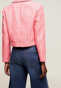 Krótka różowa kurtka z tweedu o kroju cropped, teksturowanej powierzchni i długich rękawach, w połączeniu z ciemnoniebieskimi dżinsami.