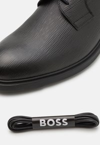 Chaussure en cuir noir avec une surface texturée, bout rond et lacets contrastants. Comprend un lacet noir avec l'étiquette "BOSS".