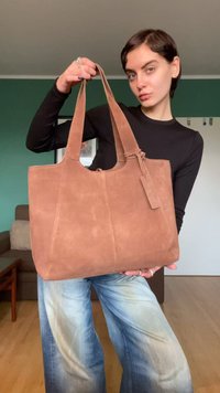 Καφέ δερμάτινη τσάντα ώμου από σουέντ με διπλές λαβές, ομαλή υφή και ορθογώνιο σχήμα. Διαθέτει ένα μικρό ταμπελάκι προσαρτημένο σε μία από τις λαβές.