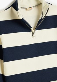 Suéter de rayas azul marino y crema con cuello con cremallera, hecho de suave material de algodón, que presenta líneas limpias y una superficie texturizada.