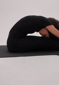 Zwarte, aansluitende leggings en een kortemouwen top op een persoon in een voorwaartse vouw yogahouding op een gestructureerde grijze mat tegen een lichte achtergrond.