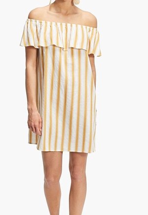 Woman wearing off-shoulder, knee-length dress with vertical yellow and white stripes and ruffled neckline, standing against white background.