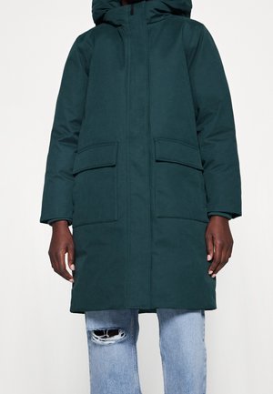 Parka à capuche vert foncé avec une fermeture éclair sur le devant, deux grandes poches à rabat et des poignets côtelés. Texture lisse, design longueur genoux et sans marquage visible.