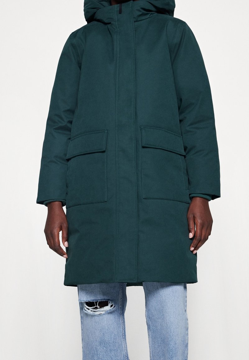 Mørkegrøn, hued parka med frontlynlås, to store påsyede lommer og ribbede mansjetter. Glat tekstur, knælangt design og ingen synlig branding.
