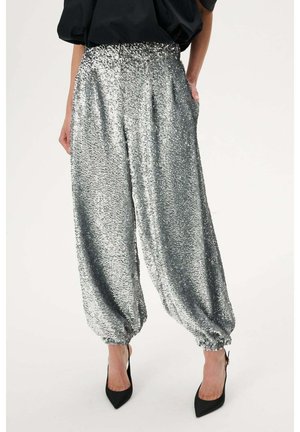 Kobieta ubrana w luźne, srebrne spodnie dresowe z cekinami z elastycznymi mankietami, w połączeniu z czarnymi szpilkami z zaostrzonym noskiem oraz czarną bluzką.